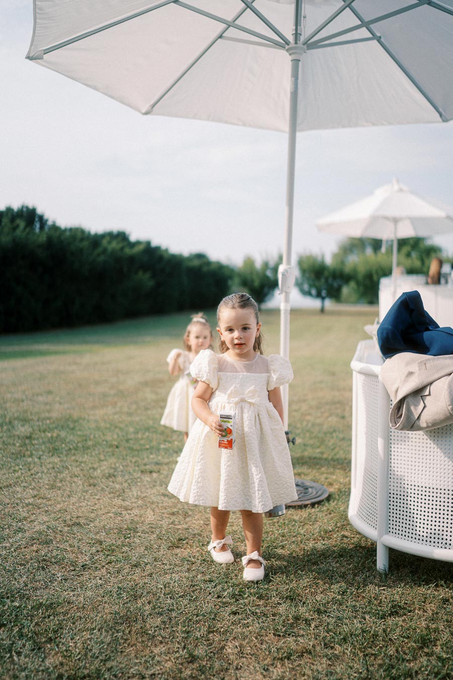 Two young girls in matching white dresses playfully walk on a grassy lawn under a large white umbrella at an outdoor event.