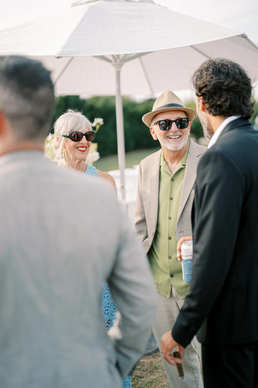 A group of well-dressed people enjoying an outdoor gathering under a large white umbrella, with a focus on a stylish older couple smiling, wearing sunglasses and hats, engaging in conversation.