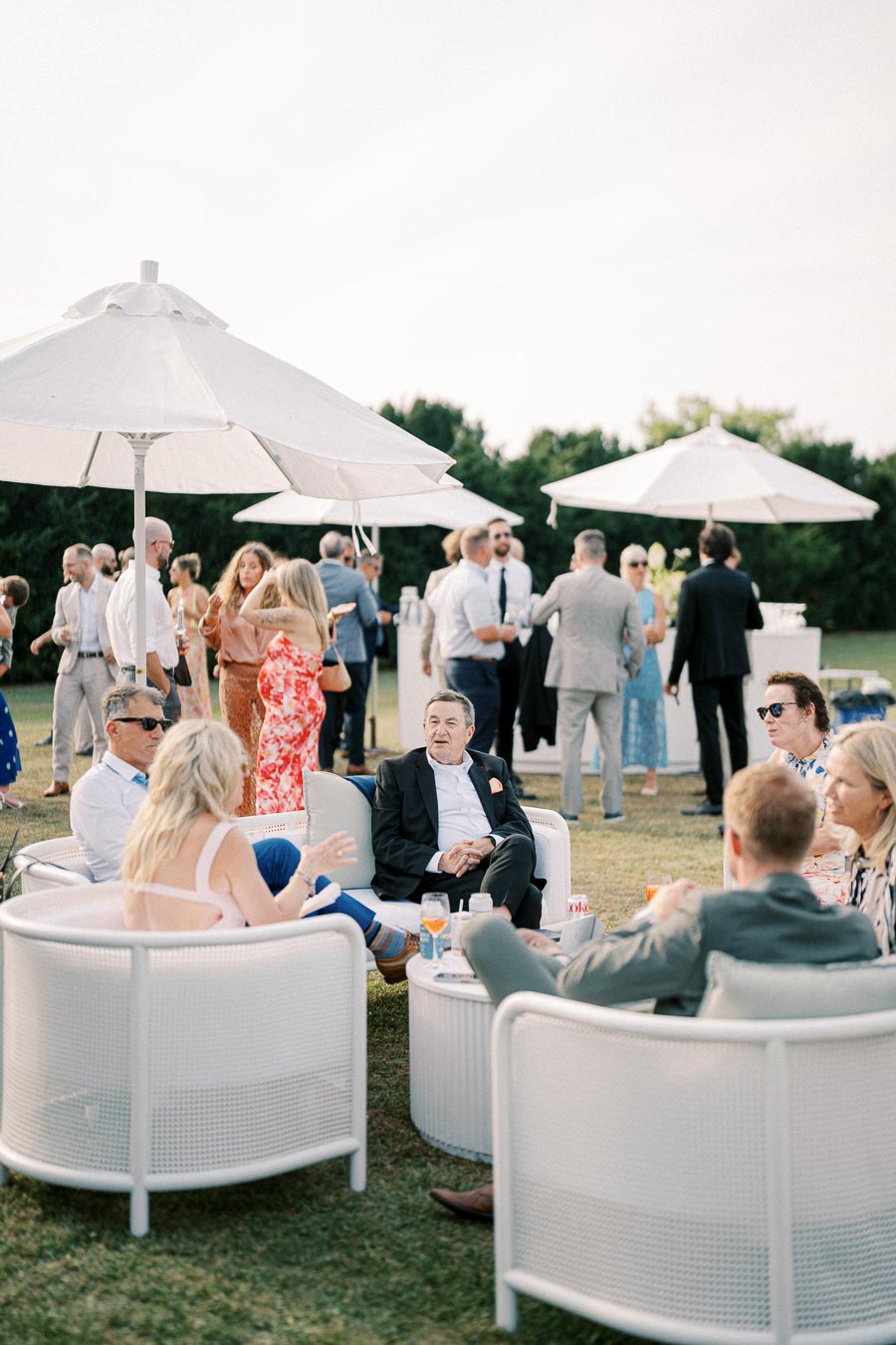 Outdoor garden party with guests socializing under white umbrellas, featuring people seated in wicker chairs and others mingling in formal attire.