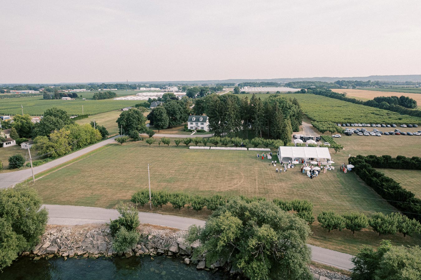 Aerial view of a countryside wedding venue with a large open field, white tent, and a historic farmhouse surrounded by lush greenery and orchards.