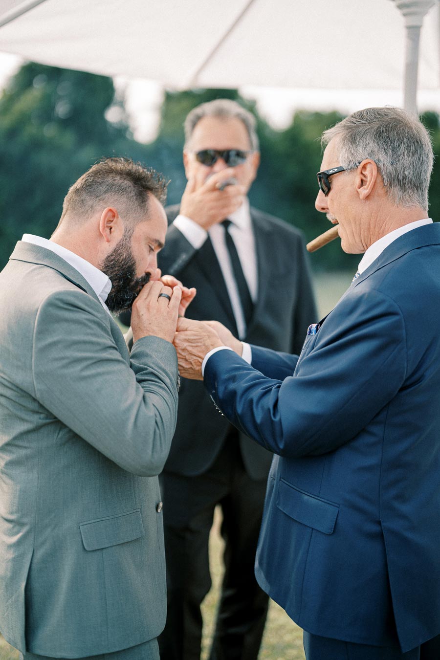 Three men in suits enjoying cigars outdoors, with one man assisting another in lighting his cigar, under a white umbrella.