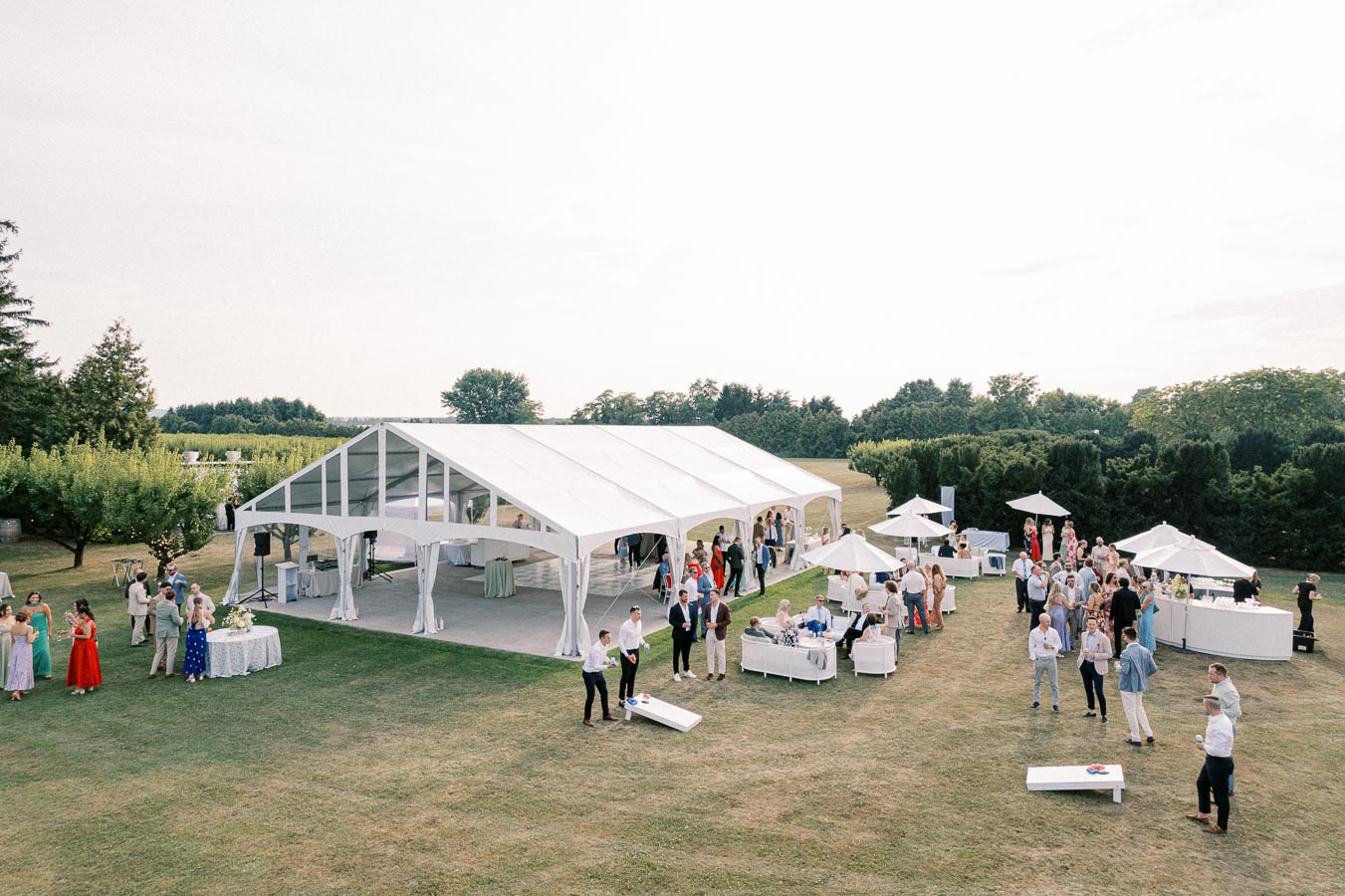 Outdoor event with a large white tent and elegant gathering of people on a grassy field, featuring white furniture and umbrellas under a clear sky.