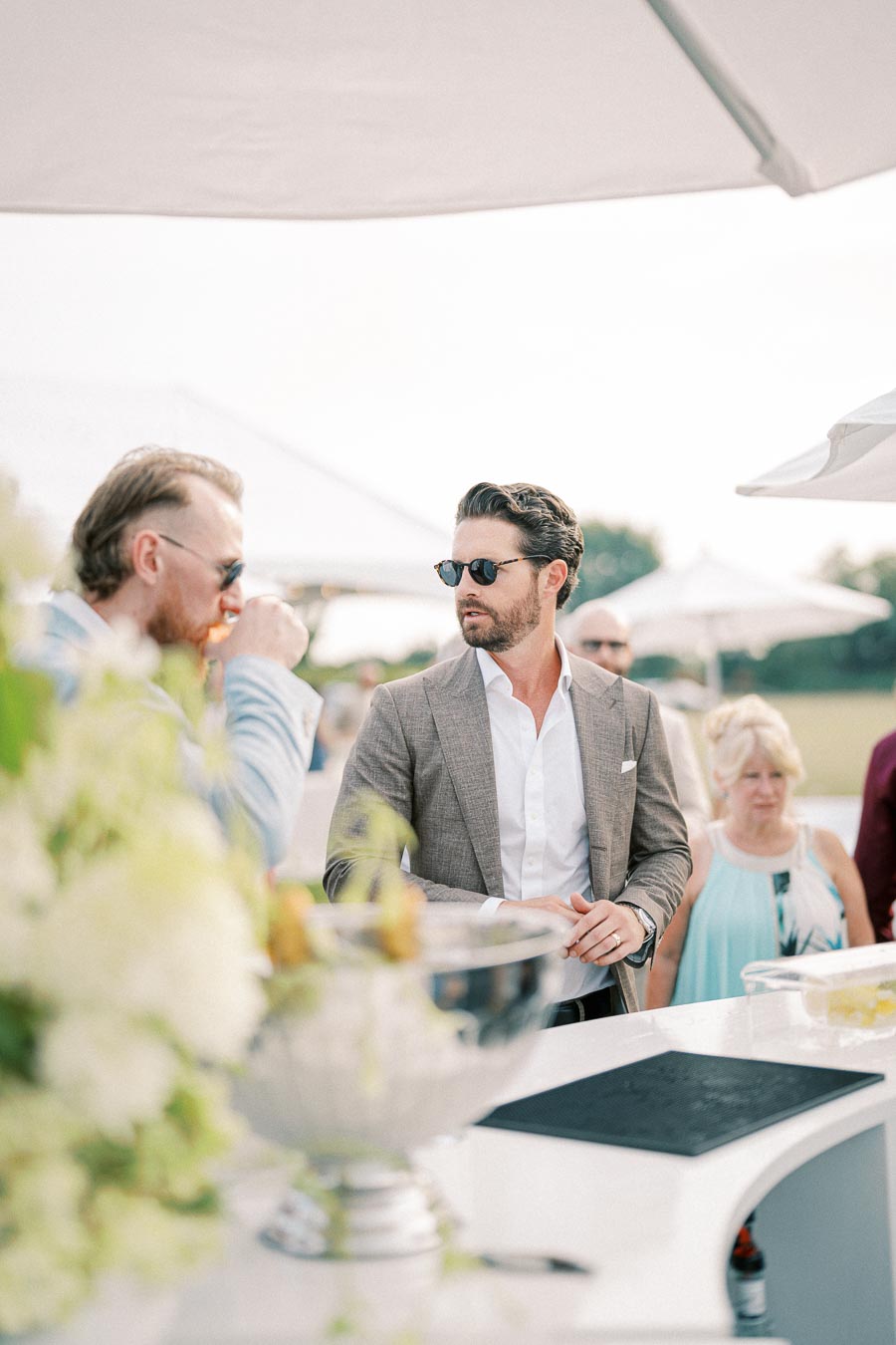 Man in a stylish suit and sunglasses attending an outdoor event, standing near a bar area with a drink, surrounded by other guests under umbrellas.