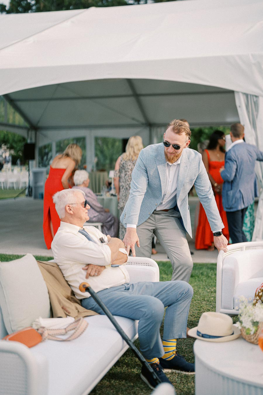 Guests at an outdoor garden party, with a man in a blue suit speaking to a seated man wearing sunglasses. A white event tent and other attendees in formal attire are visible in the background.