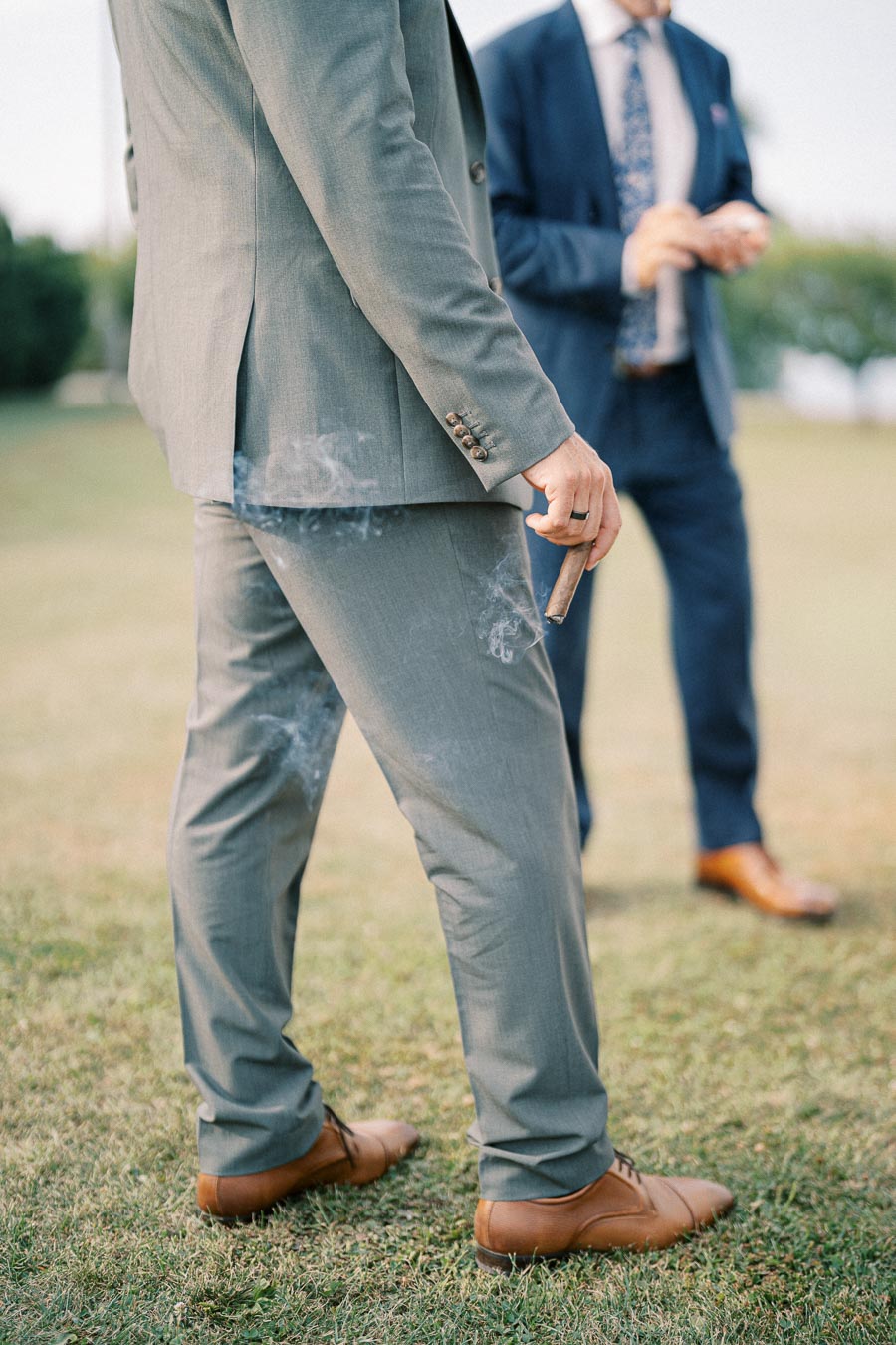 Man in a gray suit holding a cigar, standing outdoors on grass, with another man in a blue suit in the background.