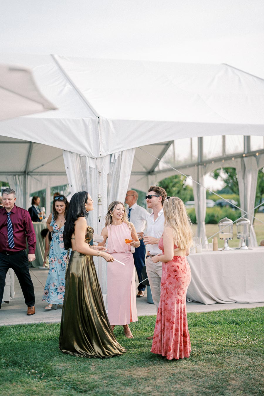 A group of elegantly dressed individuals socialize and enjoy drinks at an outdoor wedding reception under a white tent, with lush greenery in the background.