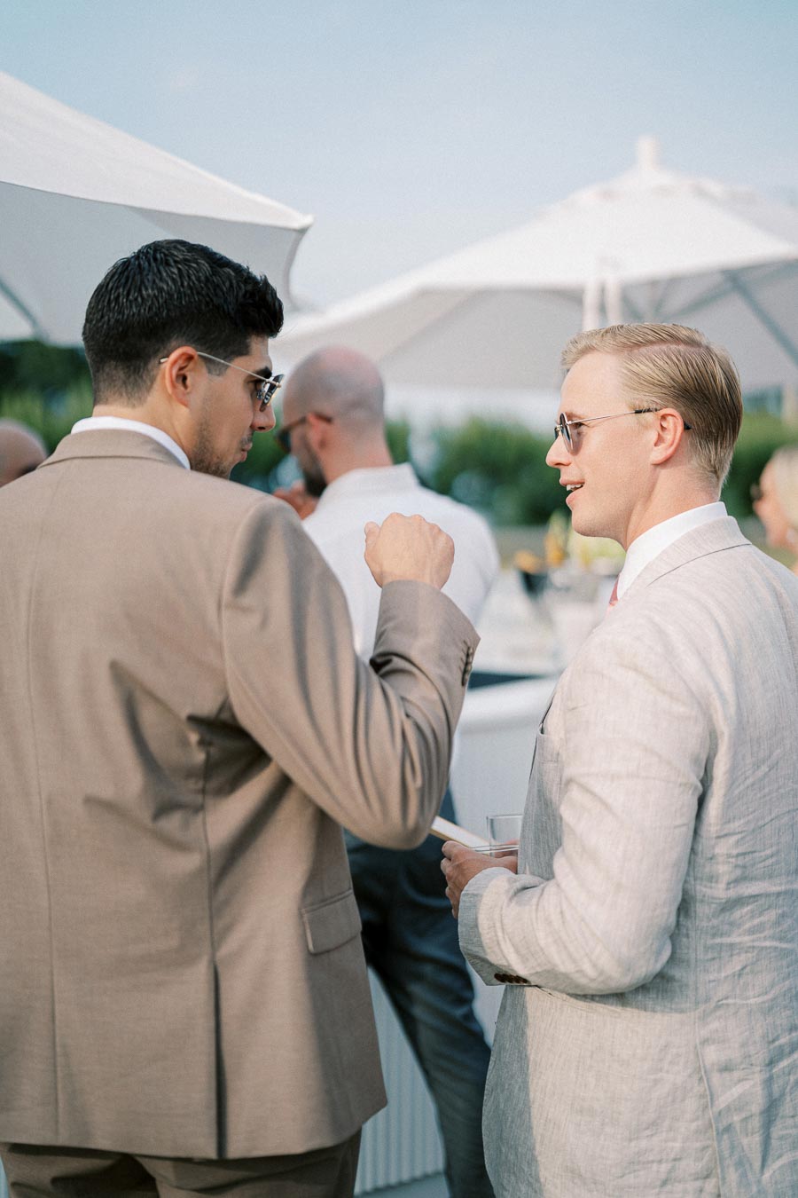 Two men in formal suits engaged in conversation at an outdoor event, surrounded by white umbrellas and greenery.