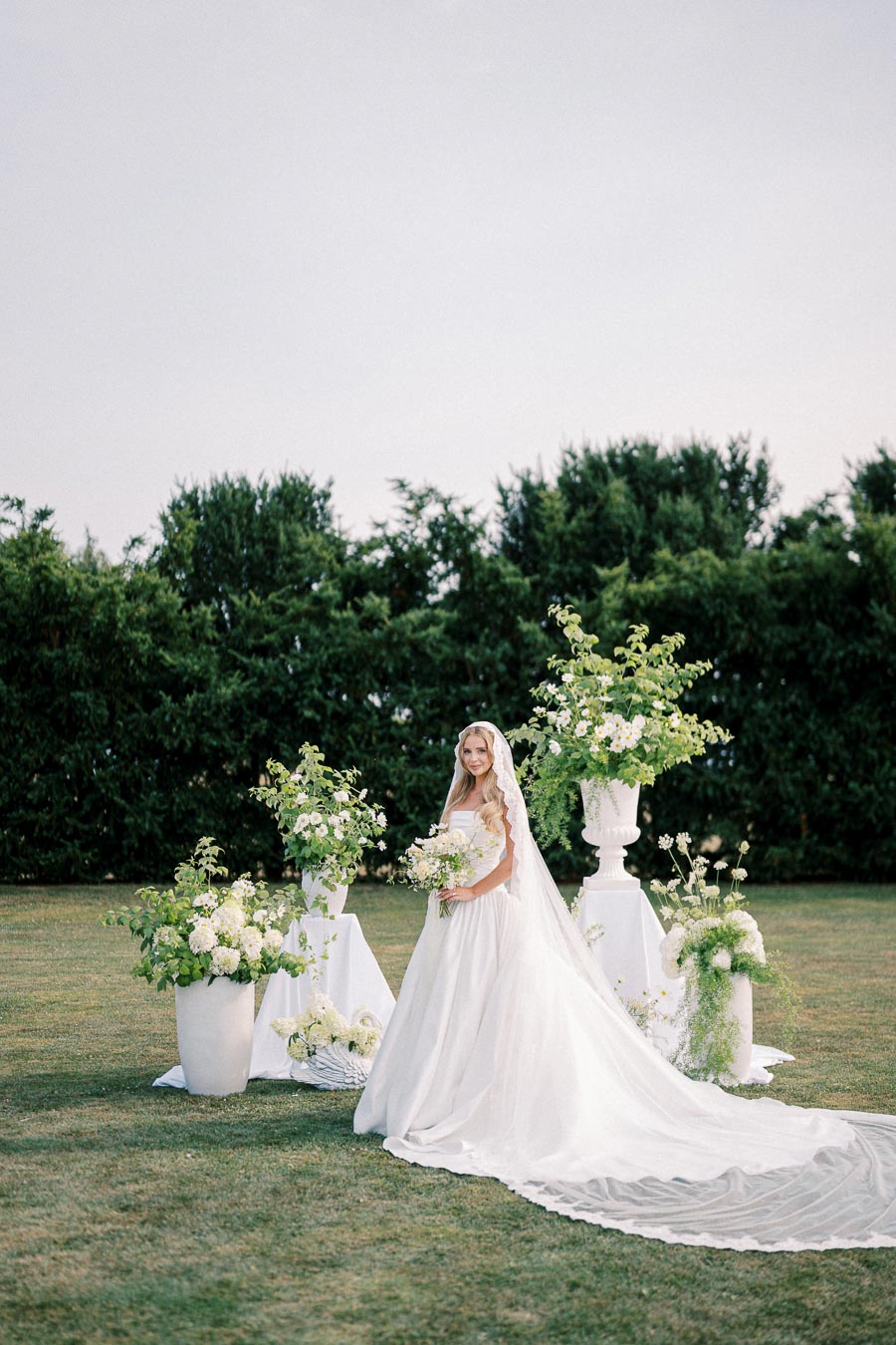 A bride in a white wedding dress and veil stands outdoors in a garden setting, surrounded by lush greenery and elegant floral arrangements.