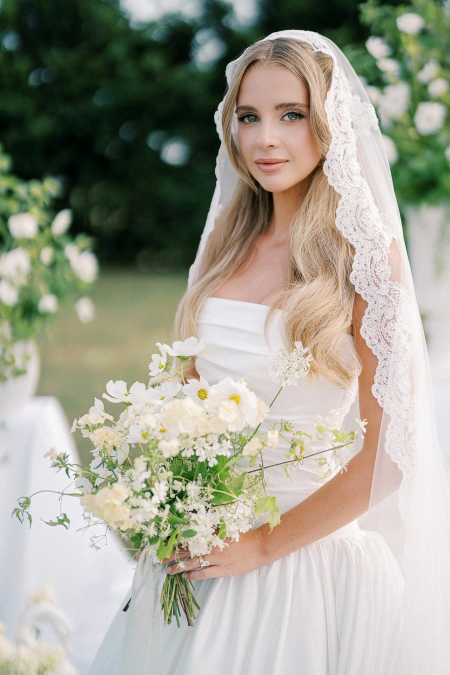 A bride in a lace veil holding a bouquet of white flowers, standing outdoors with lush greenery in the background.