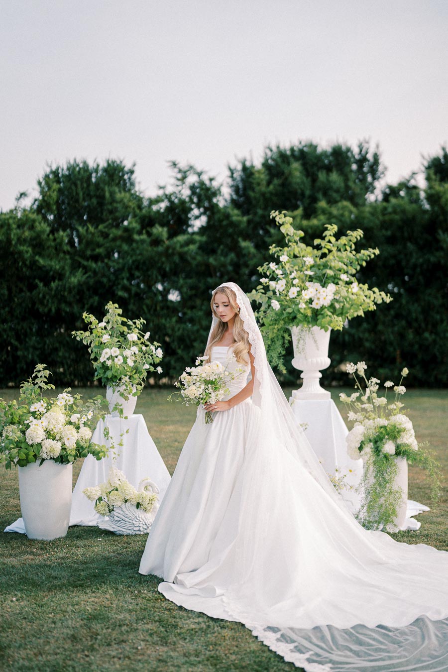 Bride in a white wedding dress with a long train, standing outdoors surrounded by lush green plants and floral arrangements, holding a bouquet of white flowers.