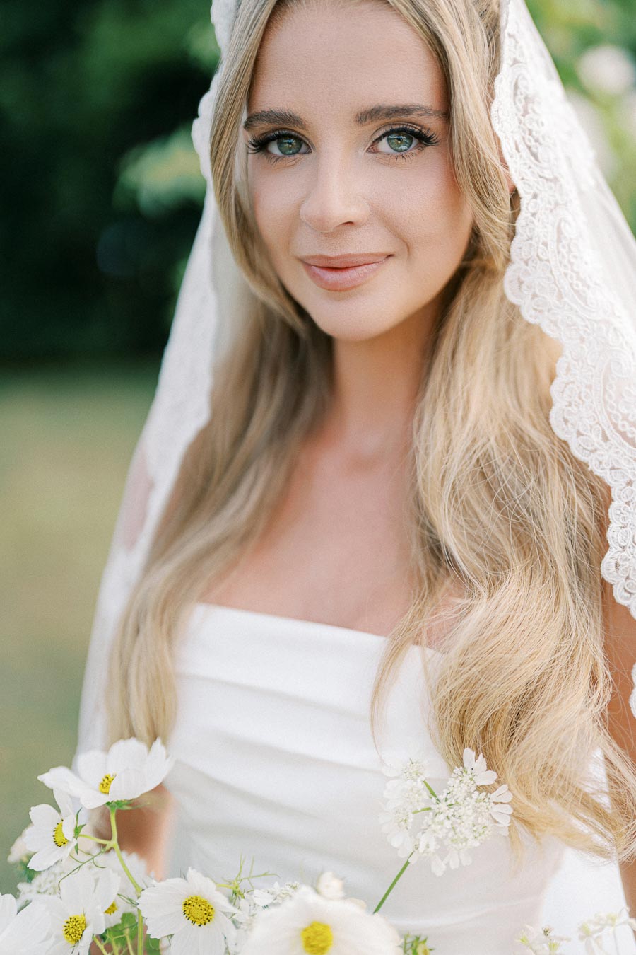 A bride with long blonde hair smiles softly, wearing a white wedding dress and a lace veil, holding a bouquet of white daisies in an outdoor setting.