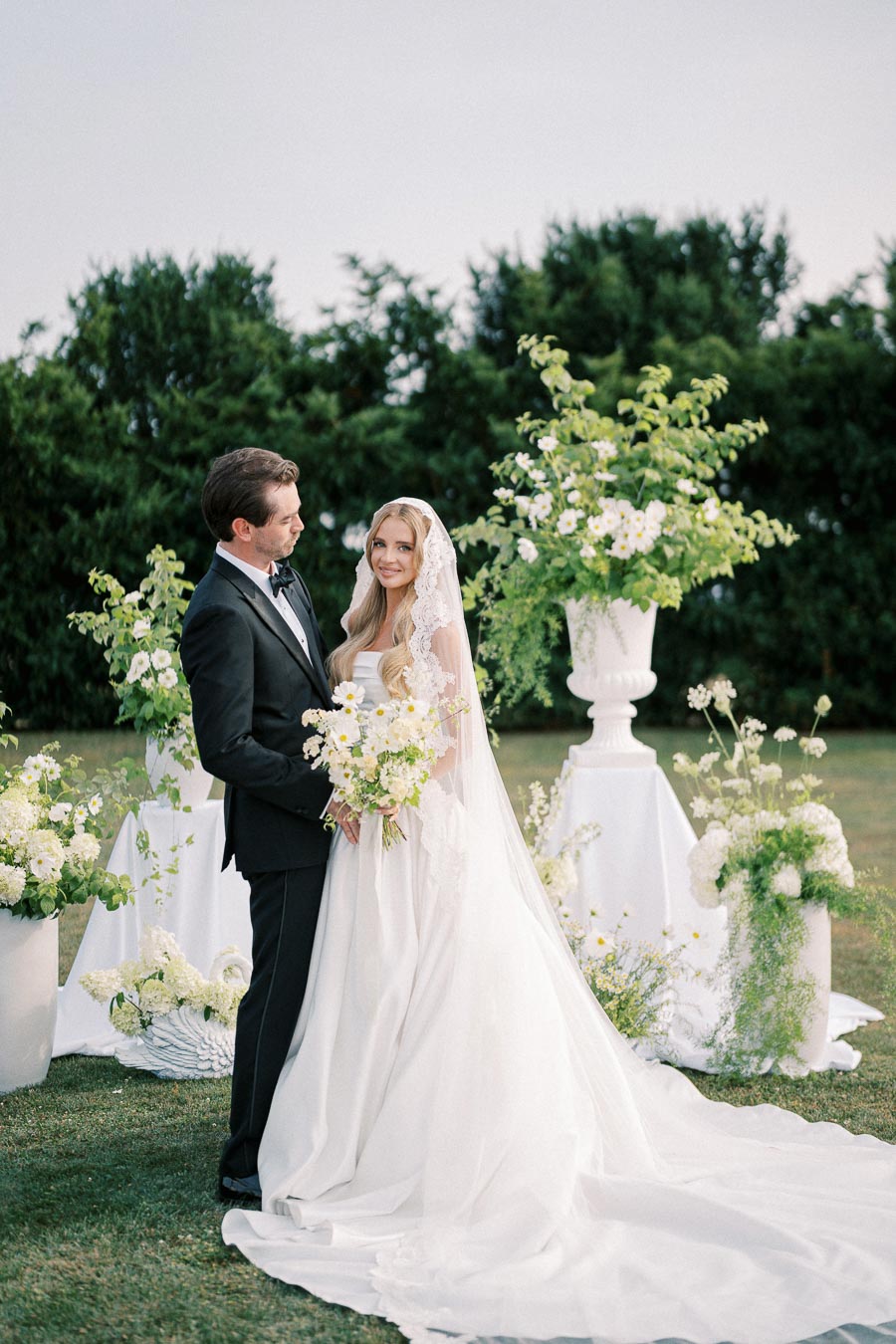 A bride and groom stand in an outdoor garden setting, surrounded by lush greenery and white floral arrangements. The bride wears a lace veil and holds a bouquet of daisies, while the groom is dressed in a classic black tuxedo. They are gazing at each other, creating a romantic wedding scene.