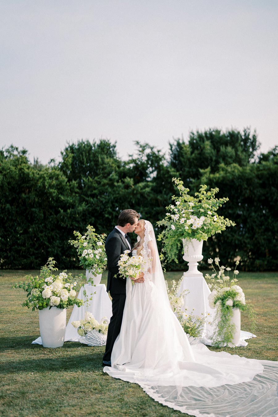 A bride and groom share a romantic kiss in an elegant outdoor wedding setting, surrounded by lush greenery and white floral arrangements. The bride wears a flowing white gown and veil, holding a bouquet, while the groom is in a classic black suit.