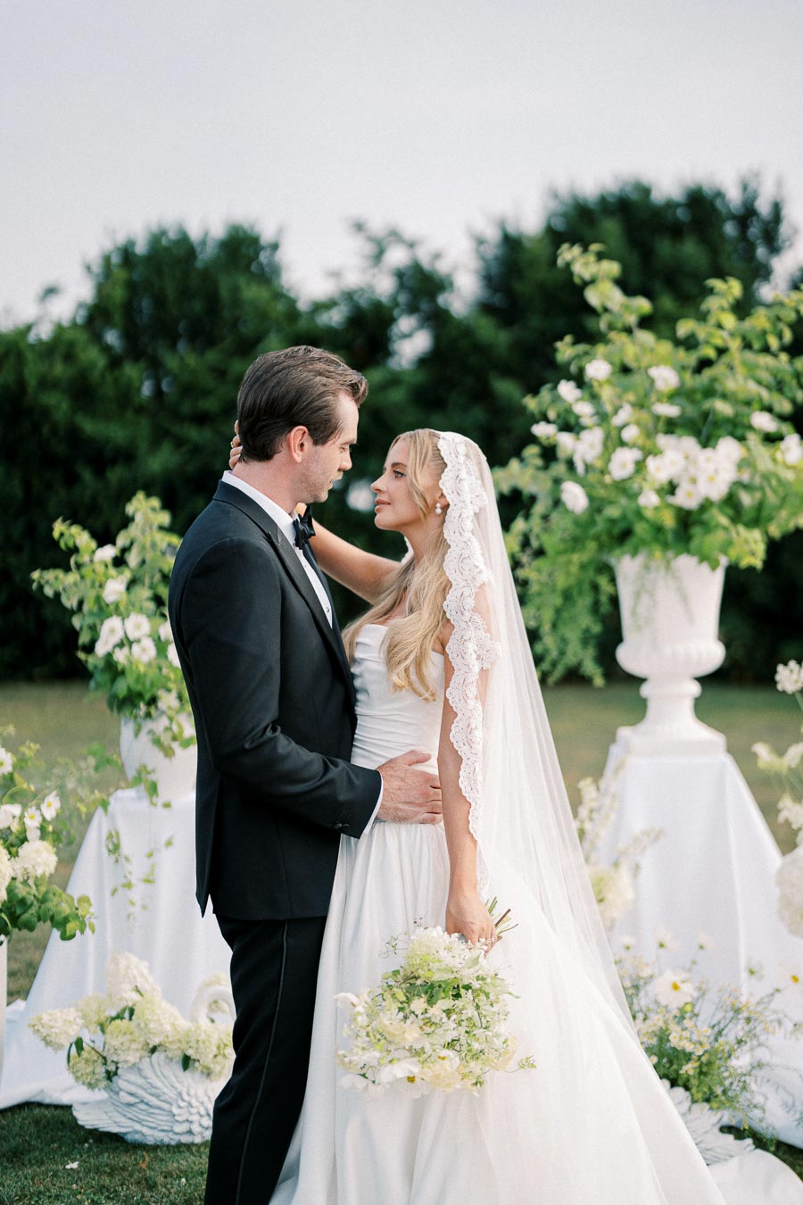 A bride and groom embracing in an outdoor garden setting, surrounded by lush greenery and white floral arrangements, with the bride wearing a lace-trimmed veil and holding a bouquet.