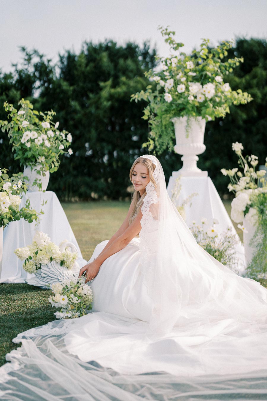 Elegant bride in a white wedding gown and veil, holding a bouquet of white flowers, seated amidst lush green floral arrangements outdoors.