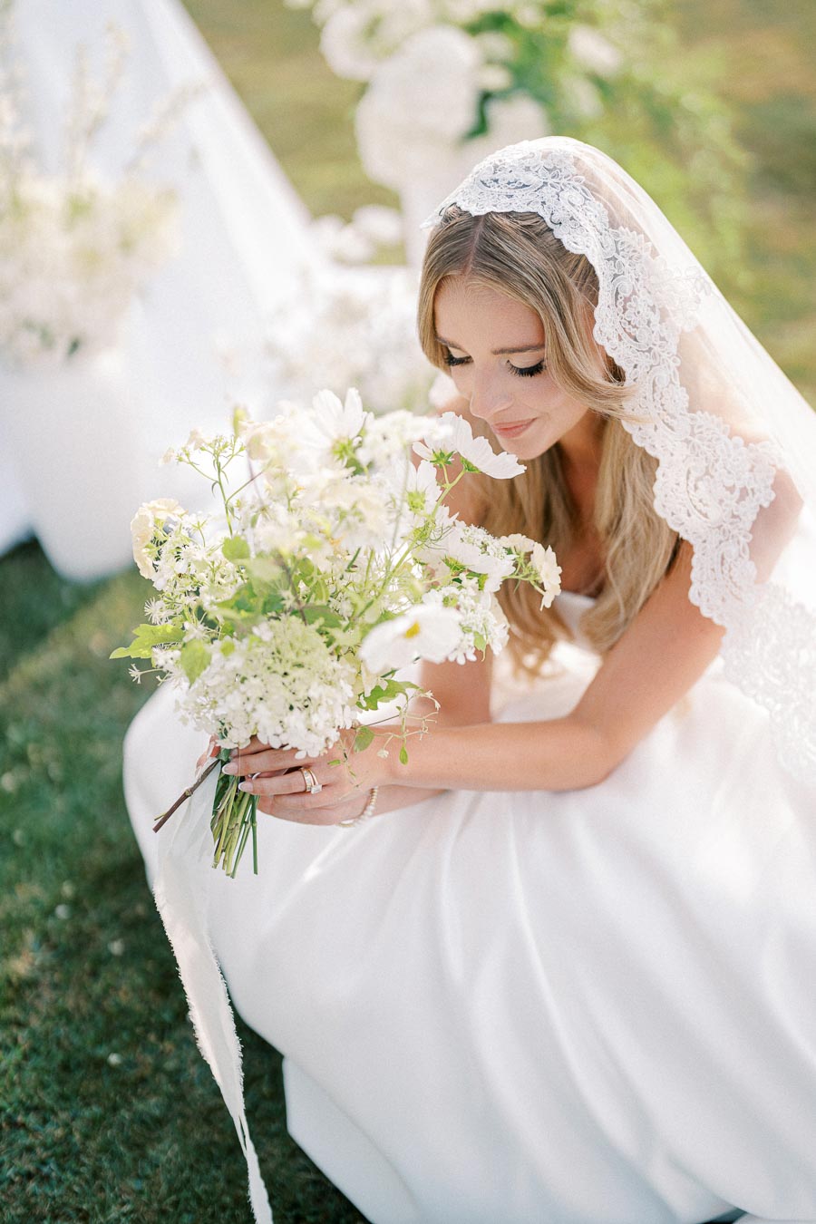 A bride with long blonde hair and a lace veil admires a bouquet of white flowers on her wedding day, surrounded by soft greenery and natural light.