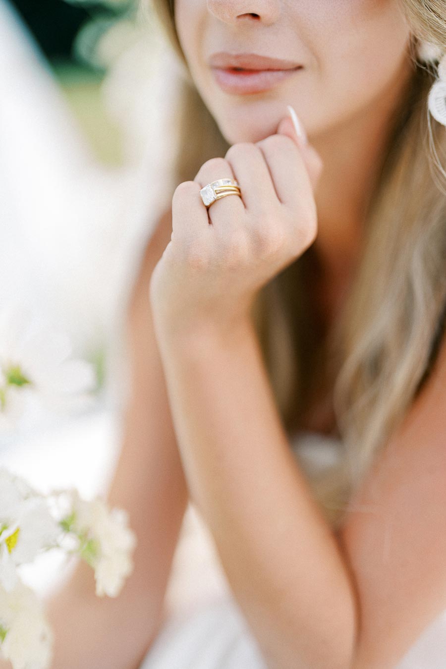 Blonde woman showcasing elegant gold engagement ring with large diamond, surrounded by soft floral background.