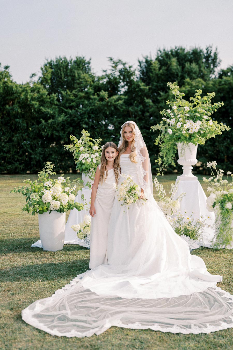 Bridal portrait outdoors with a young girl, featuring a bride in a white wedding dress and lace veil, holding a bouquet of white flowers, standing beside lush greenery and floral arrangements on a sunny day.