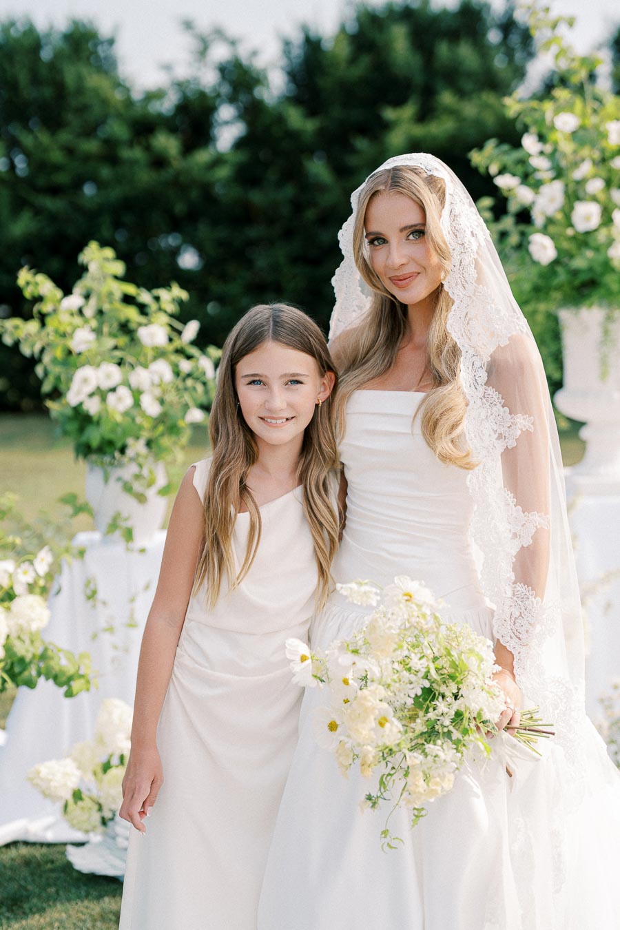 Bride in elegant white gown and lace veil posing with young girl at an outdoor wedding ceremony, surrounded by lush greenery and white floral arrangements.