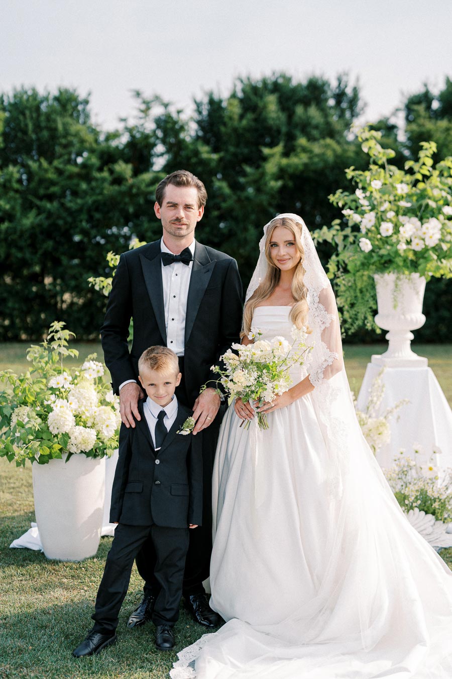 A bride and groom, dressed in formal attire, pose with a young ring bearer in an outdoor wedding setting surrounded by lush greenery and floral arrangements.