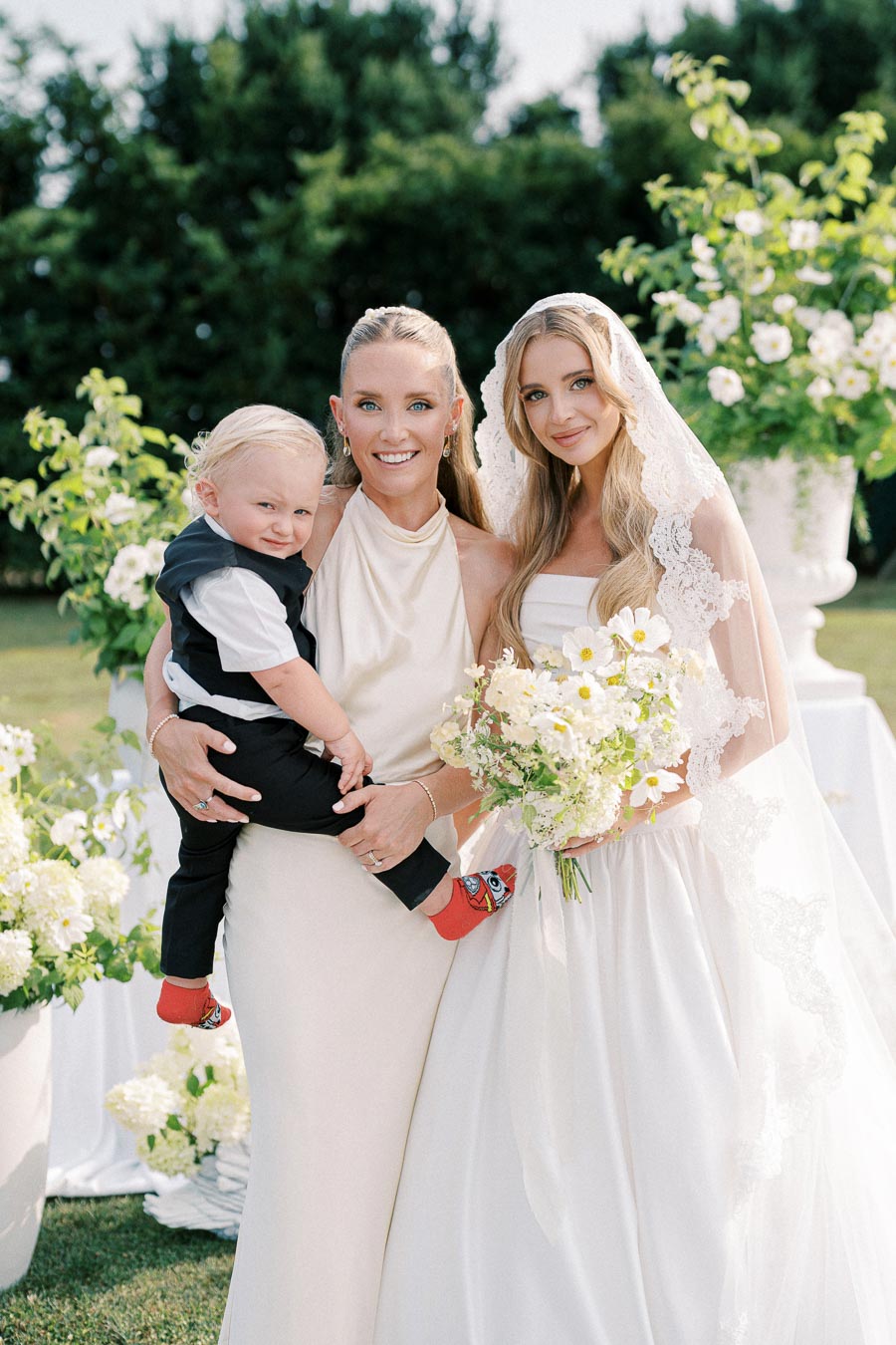 A joyful wedding scene featuring a bride in an elegant white gown and lace veil holding a bouquet of flowers, accompanied by a smiling woman in a cream dress holding a child dressed in a vest and red socks, surrounded by lush greenery and white blossoms.