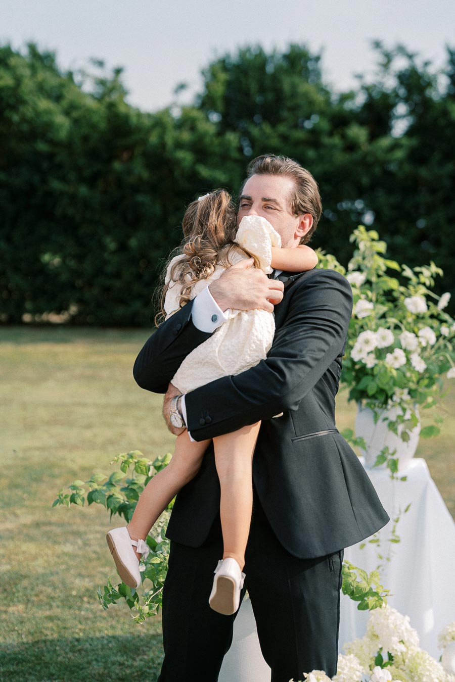 A man in a suit joyfully embraces a young girl in a white dress, outdoors with greenery and flowers in the background.