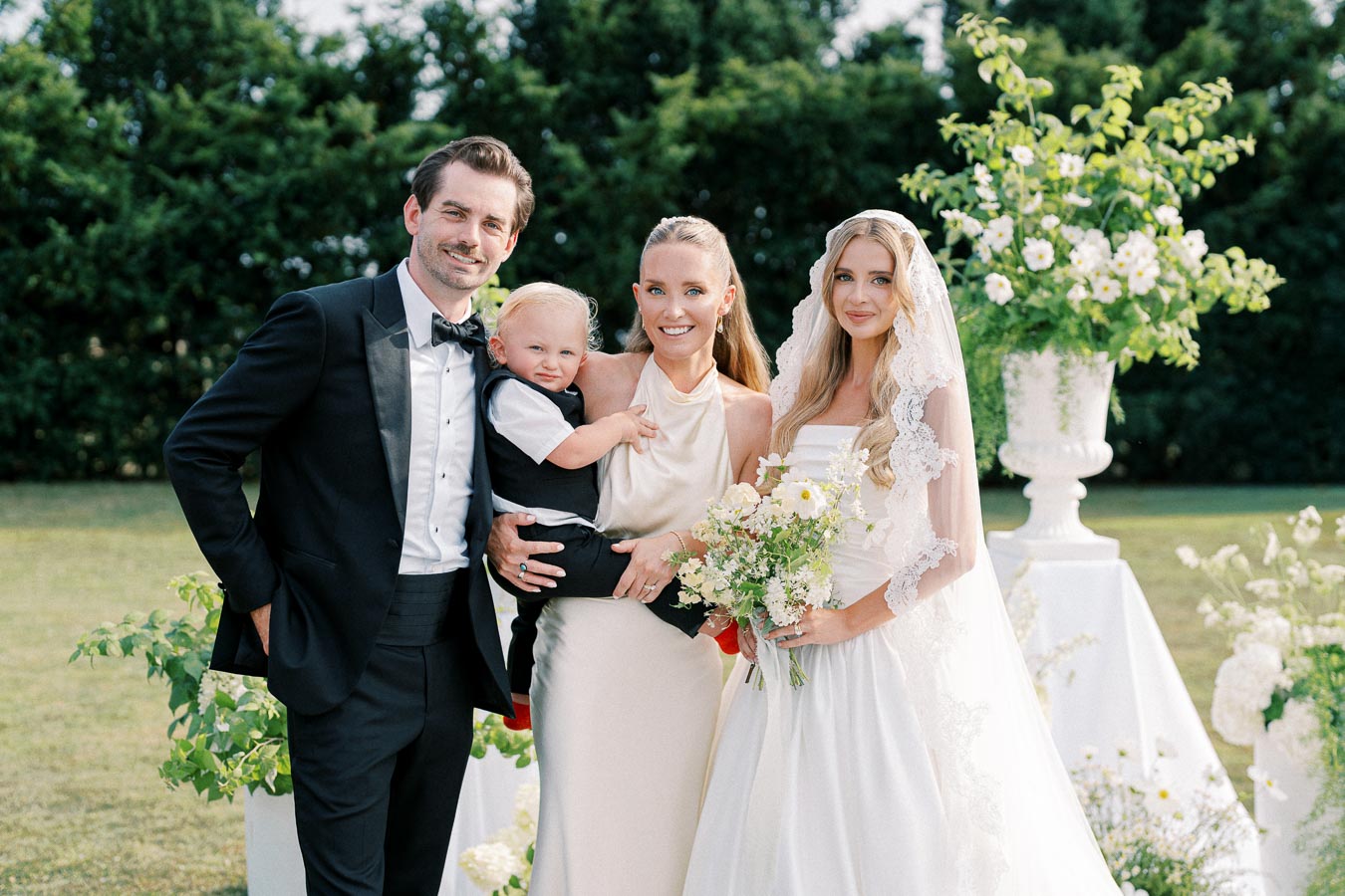 A happy couple poses with a family in elegant wedding attire, surrounded by lush greenery and floral arrangements, capturing a joyful outdoor wedding ceremony.