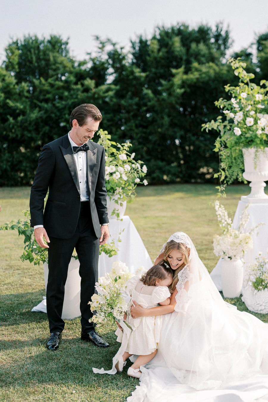 Bride in a white wedding dress smiling and hugging a small child during an outdoor wedding ceremony, with the groom in a black tuxedo looking on and green foliage in the background.