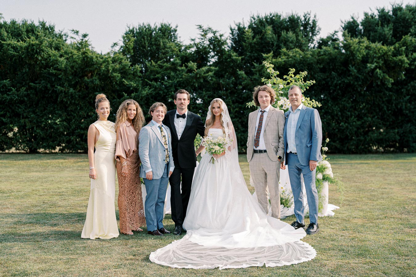 A wedding party posing outdoors with the bride in a white gown, holding a bouquet, flanked by the groom in a black tuxedo, and five guests in formal attire, standing on a green lawn with lush trees in the background.