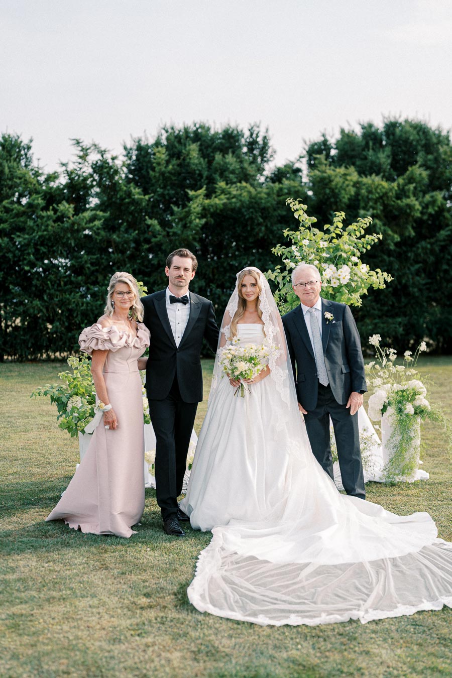 Wedding party standing outdoors with a bride in a white gown and veil holding a bouquet, flanked by three elegantly dressed people in formal attire, with lush greenery in the background.