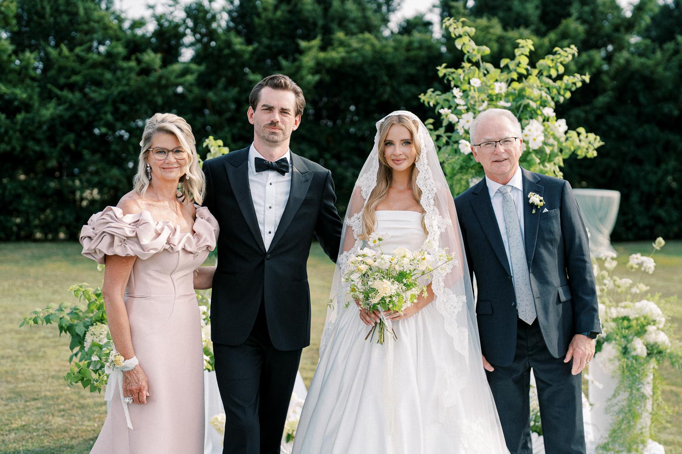 A bride in a white gown and lace veil stands with a groom in a black tuxedo, alongside an elegantly dressed woman in a pink gown and a man in a suit. They are outdoors surrounded by greenery and flowers, capturing a joyous wedding moment.