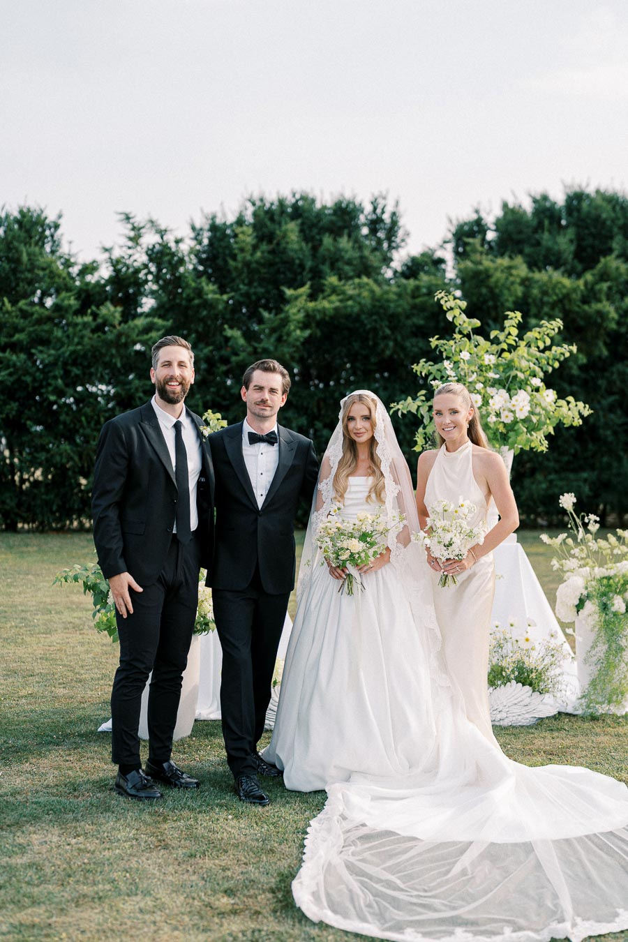 A bride and groom pose with two guests at an outdoor wedding ceremony. The bride wears a white gown with a lace veil, the groom and one guest are in black suits, while the other guest wears a cream dress. They are surrounded by floral arrangements and greenery.