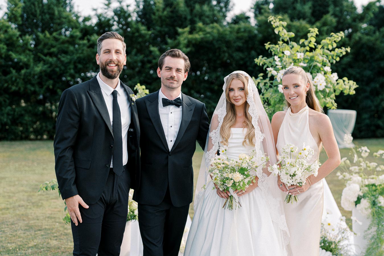 A joyful wedding scene with two groomsmen in black tuxedos and two bridesmaids in white dresses, standing outdoors surrounded by greenery and flowers. The bride in a traditional wedding gown with a lace veil holds a bouquet of white and yellow flowers, creating an elegant and serene atmosphere.
