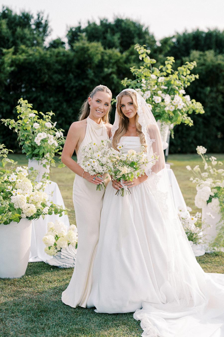 Two women standing outdoors in elegant attire, one in a wedding gown and veil, the other in a light-colored dress, both holding bouquets of white flowers, surrounded by lush greenery and floral arrangements.