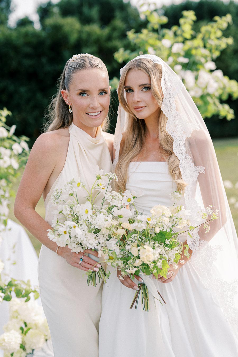 Two women in elegant dresses, one in a white wedding gown with a lace veil, holding bouquets of white and green flowers in a sunny garden setting.