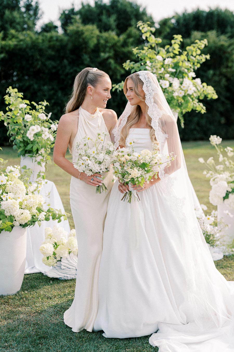 Two women in elegant white dresses, one in a halter gown and the other in a bridal gown with a lace veil, smiling at each other while holding flower bouquets in a lush garden setting.