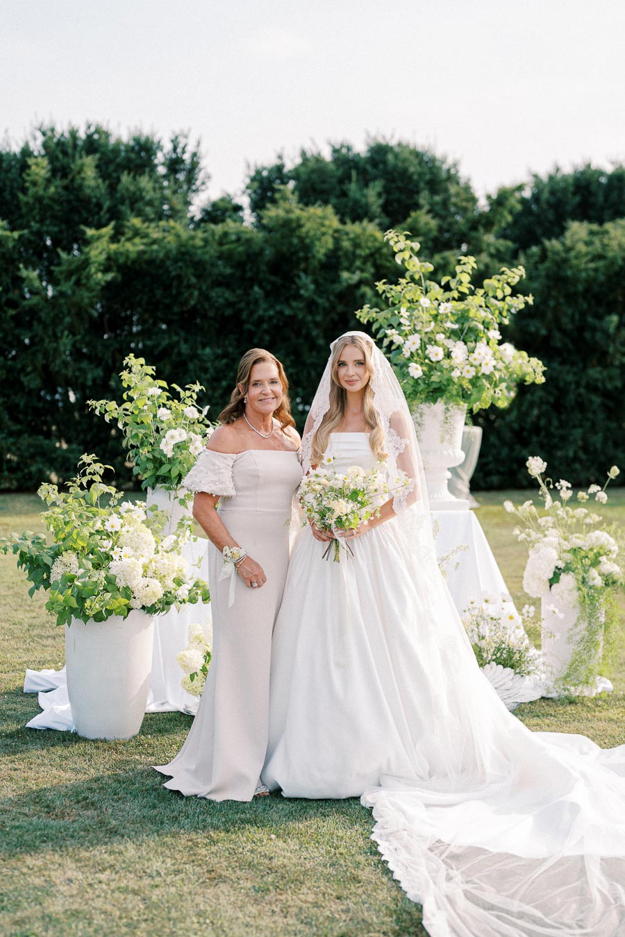 A bride in a flowing white gown stands next to a woman in a light pink dress, surrounded by lush greenery and white floral arrangements in an outdoor garden setting.