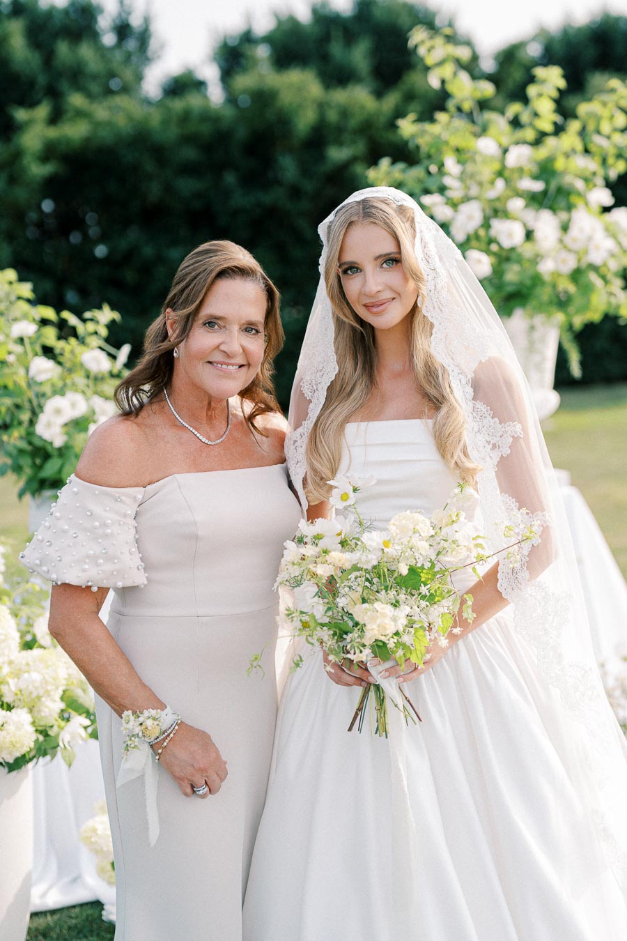 Smiling bride wearing a lace veil and holding a bouquet, standing with a woman in an off-shoulder dress adorned with pearls, both surrounded by white flowers and greenery outdoors on a sunny day.