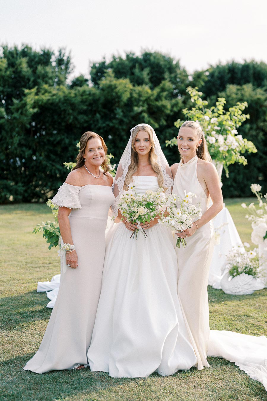 Three women smiling at an outdoor wedding; the bride in a white gown and lace veil holds a bouquet, flanked by two women in elegant dresses holding flowers, with lush greenery in the background.