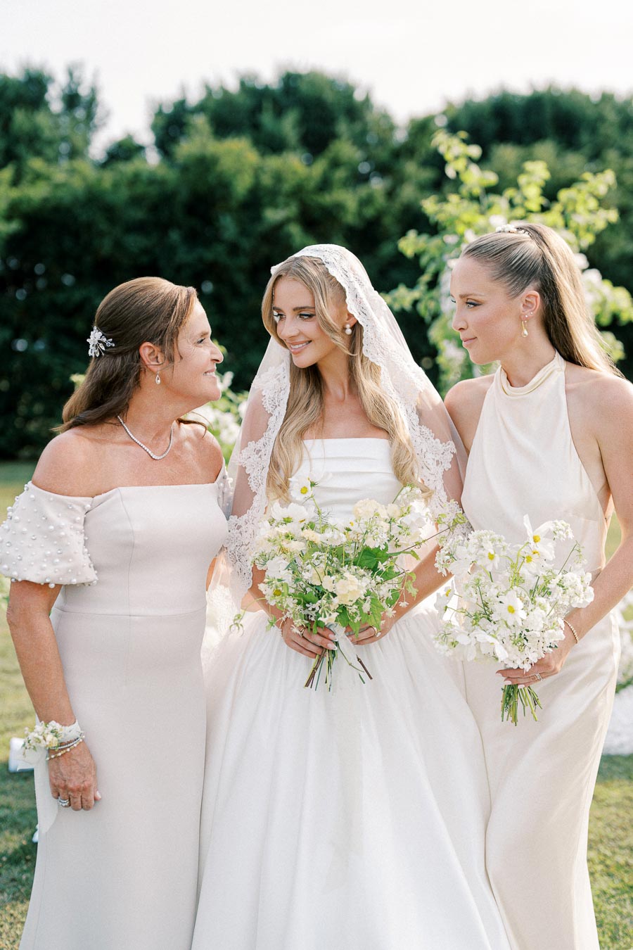 Bride in elegant white dress and lace veil stands smiling with two women holding floral bouquets against a lush garden backdrop, capturing a joyful wedding moment.