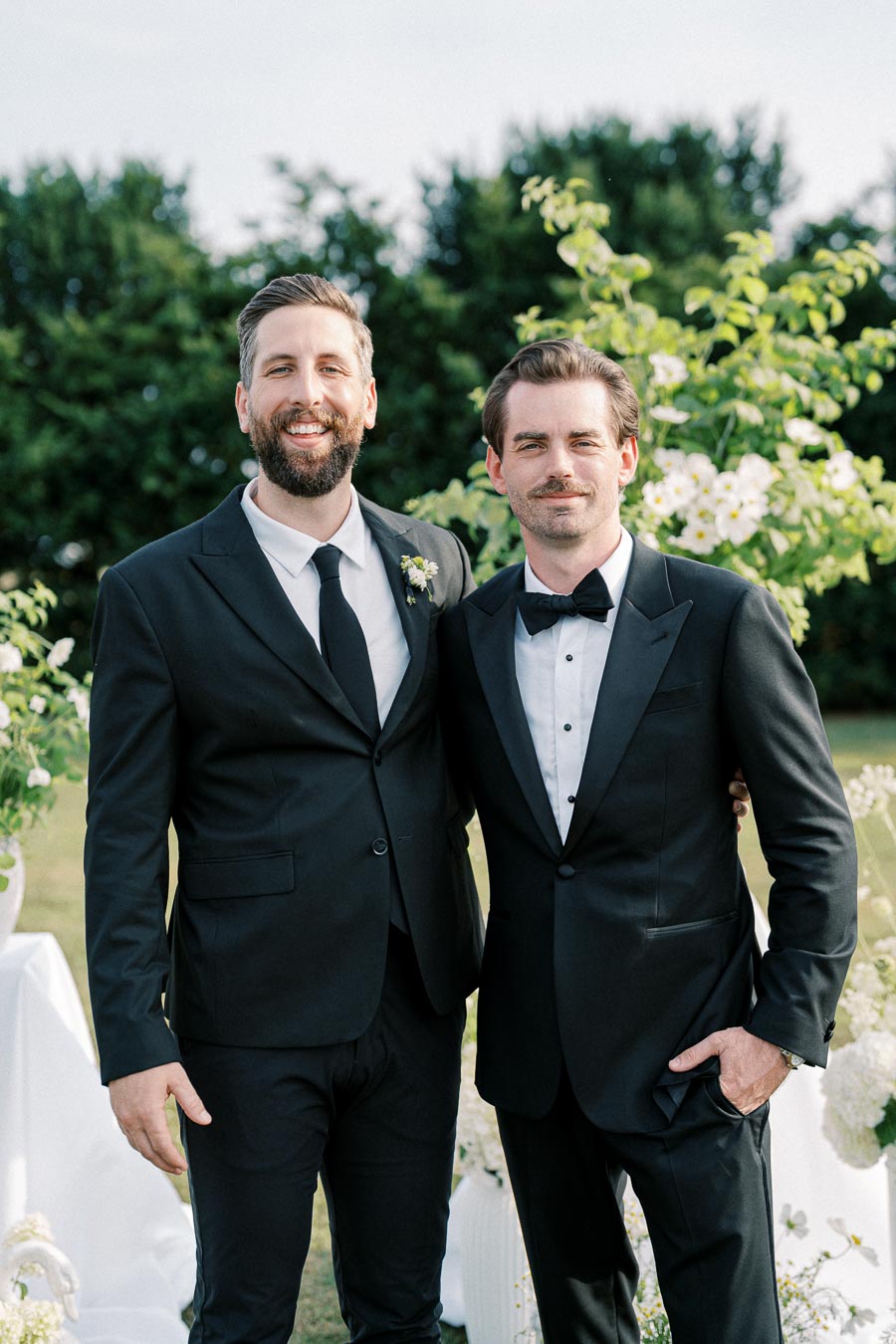 Two men in formal black suits and ties pose outdoors with green foliage and white flowers in the background, possibly at a wedding or formal event, smiling warmly at the camera.