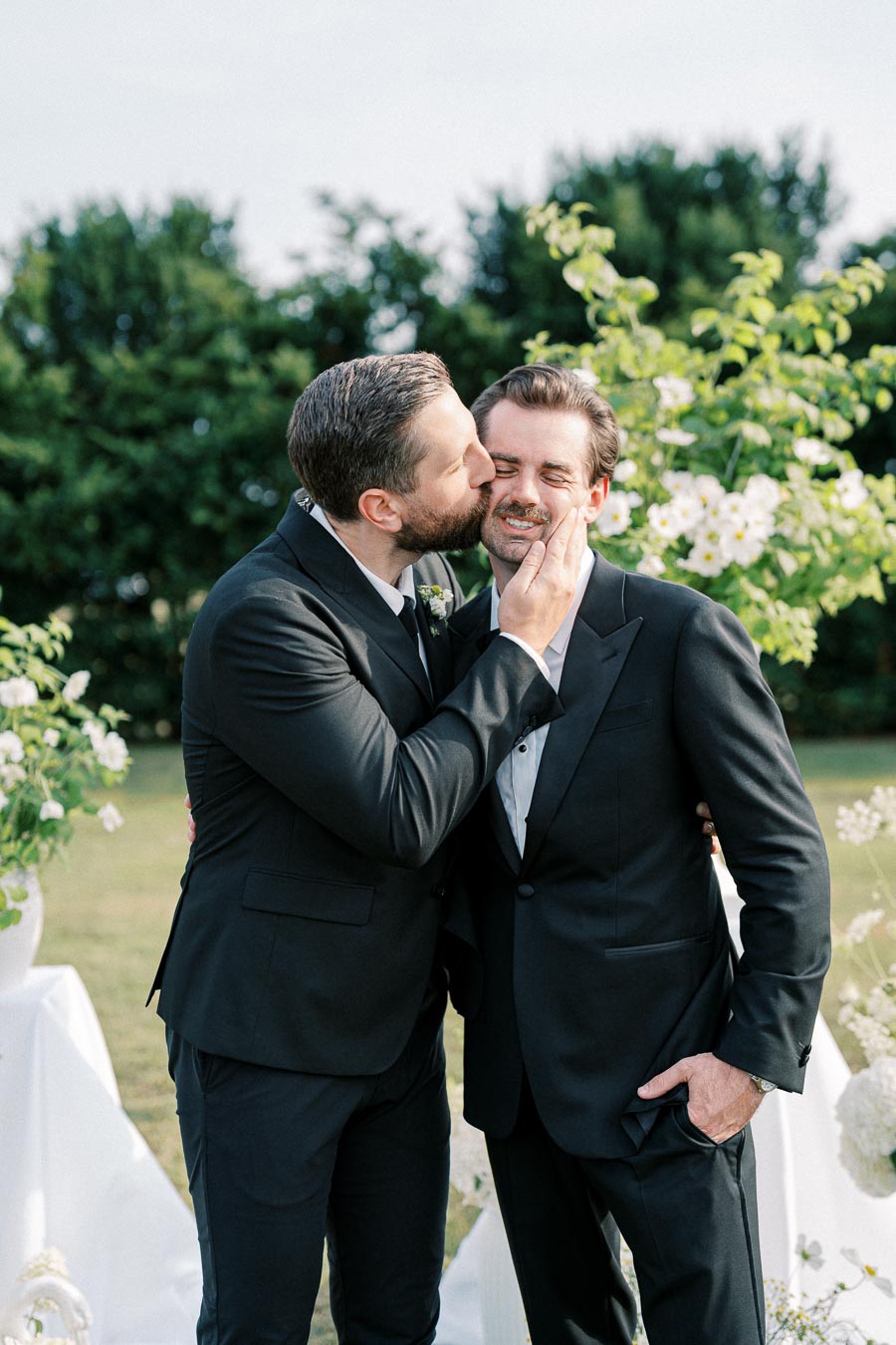 Wedding celebration featuring two men in suits, sharing a joyful moment with a kiss on the cheek amidst lush greenery and floral decor.