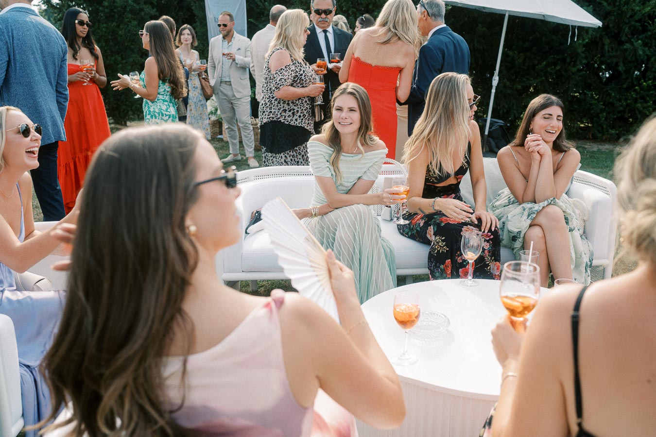 Group of elegantly dressed people enjoying an outdoor social gathering, seated around a white table with drinks, surrounded by lush greenery.