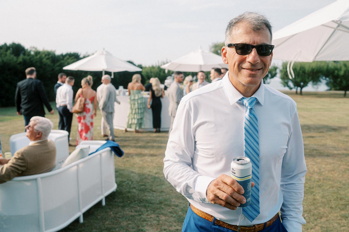 Man in sunglasses holding a drink at an outdoor social event, with elegantly dressed people mingling in the background under white parasols on a grassy field.