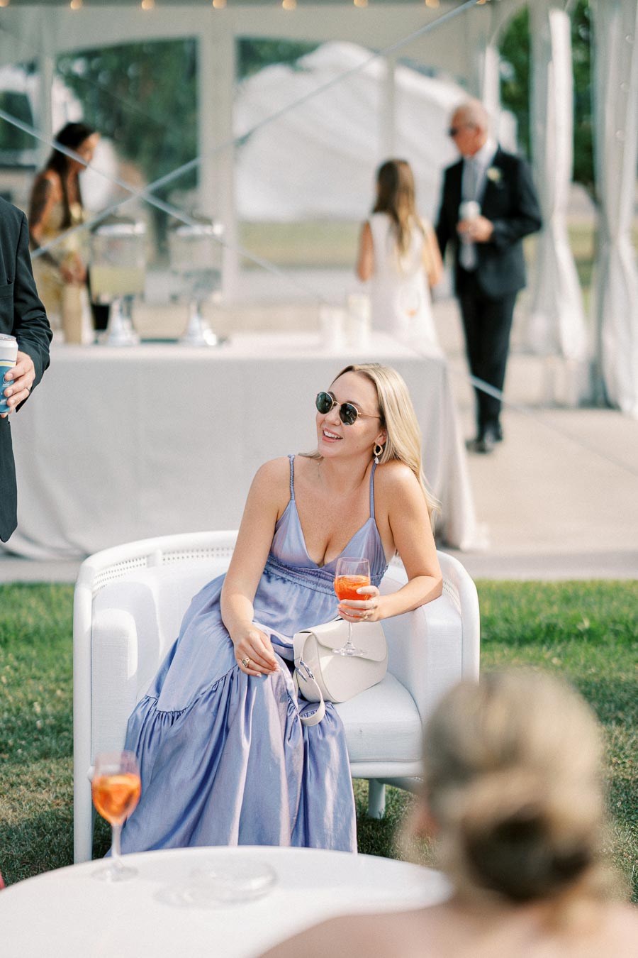 A woman in a light blue dress and sunglasses sits on a white chair at an outdoor event, holding a glass with an orange drink. The scene includes other attendees in the background and a white canopy tent.