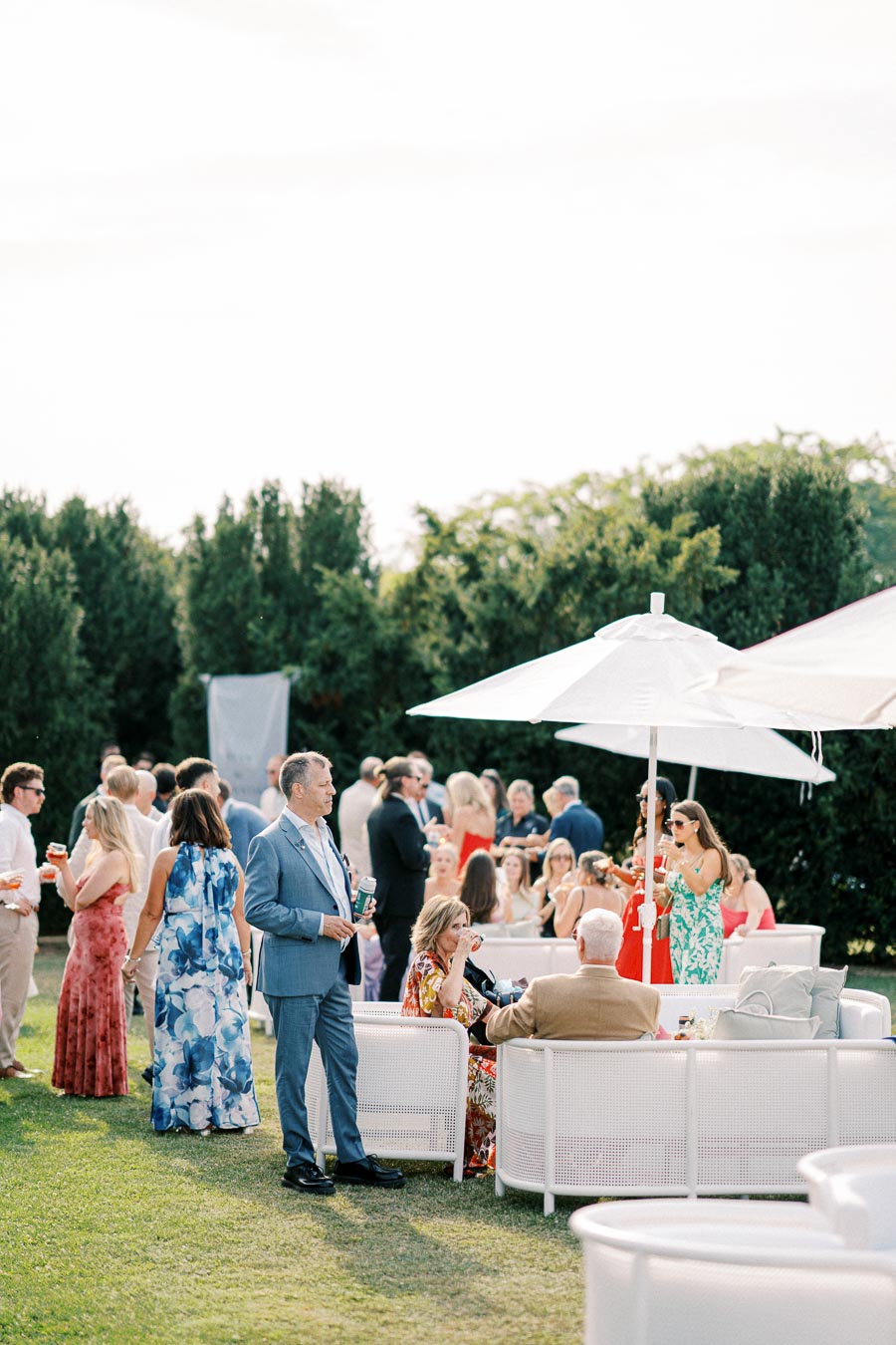 Elegant outdoor garden party with guests socializing under a white umbrella, surrounded by lush greenery, dressed in colorful summer attire.