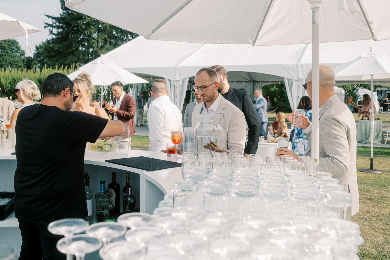 People enjoying drinks at an outdoor wedding reception under white tents, with a bartender serving beverages and guests socializing in the background.