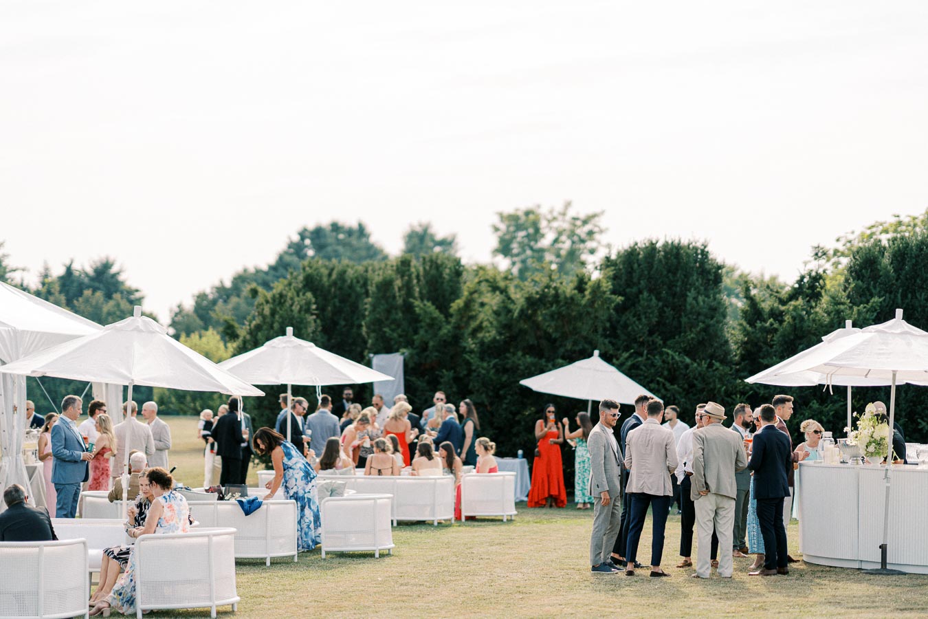 Outdoor garden party with elegantly dressed guests socializing under white umbrellas, featuring modern seating areas and lush greenery in the background.