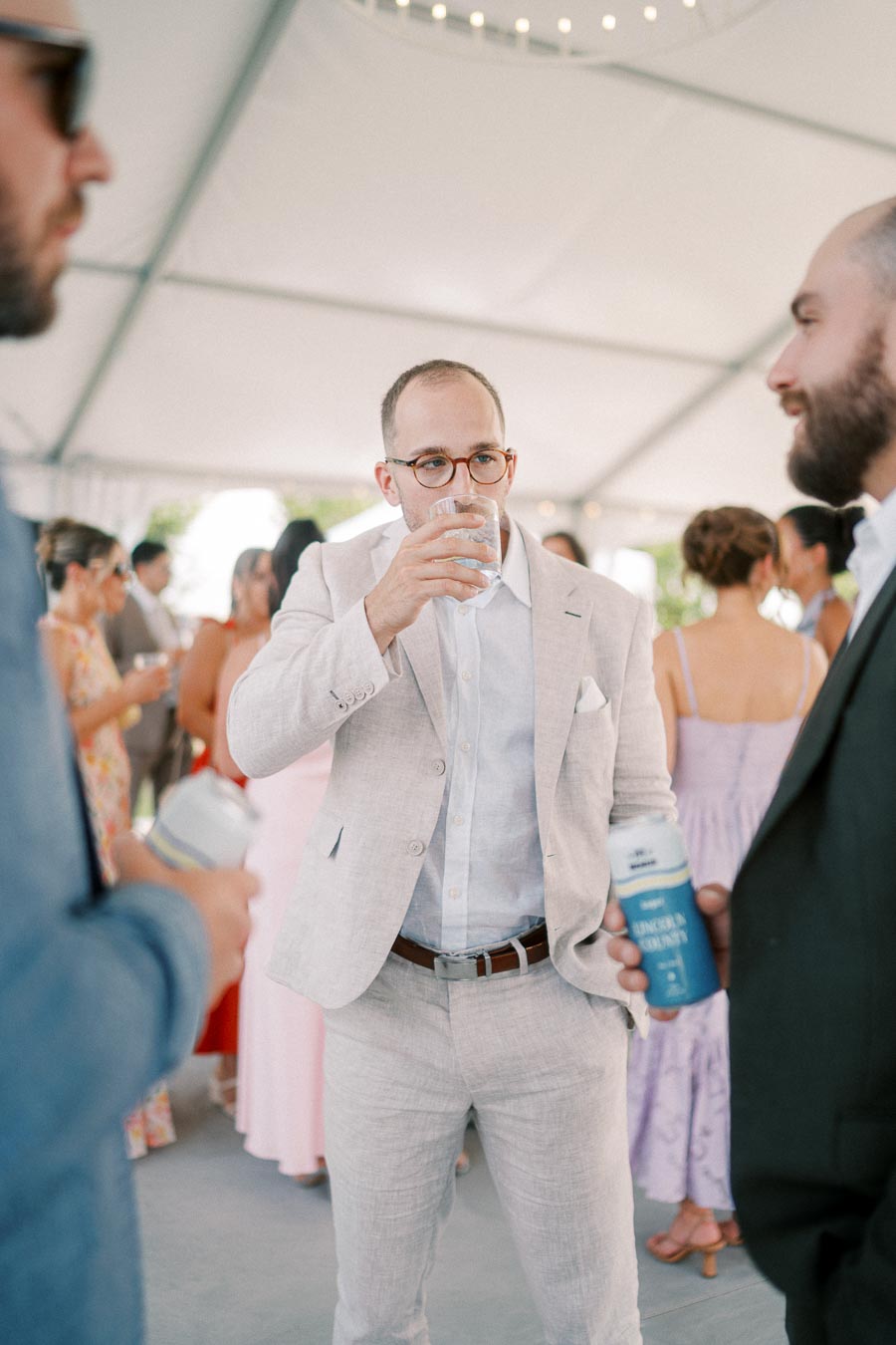 Man in a beige suit drinking water at a social gathering with people mingling under a canopy.