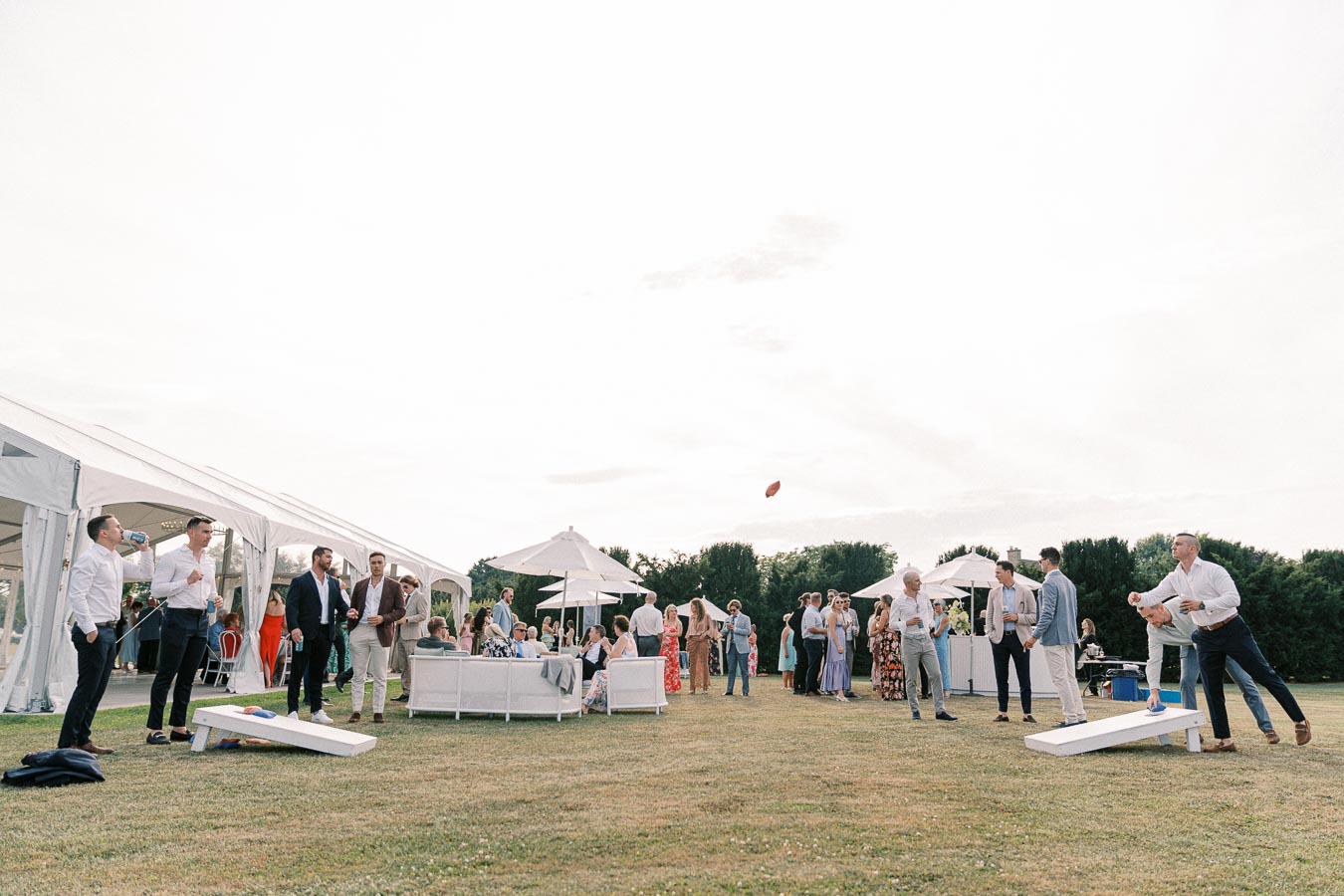 Outdoor wedding reception with guests playing lawn games, socializing under a white marquee, and enjoying a sunny day on a grassy field.